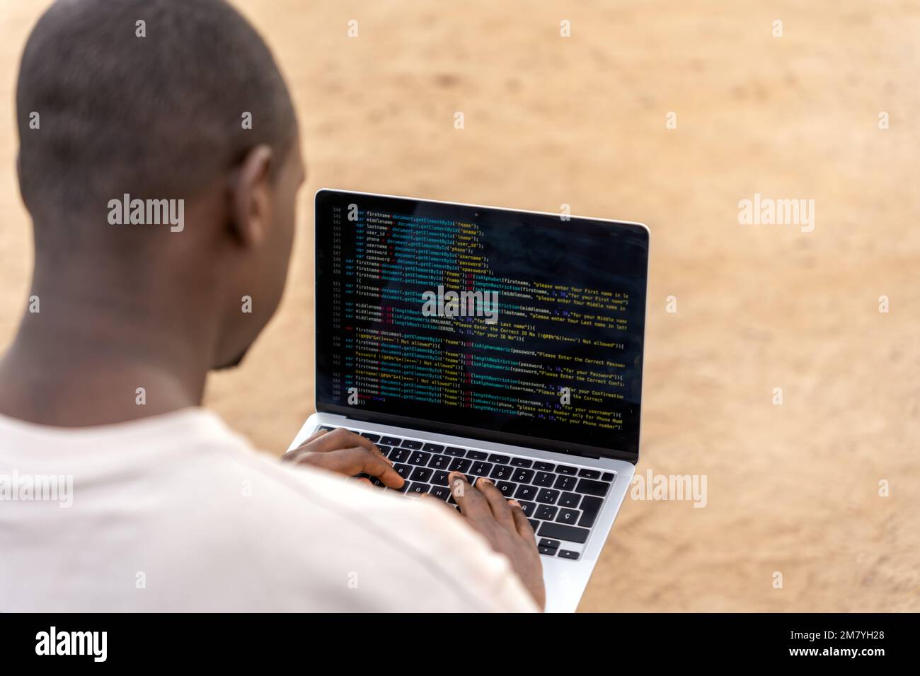 Ein Afroamerikaner in lässiger Kleidung, der seinen Laptop benutzt, um Code für Software zu erstellen, während er Remote am Strand arbeitet Stockfoto