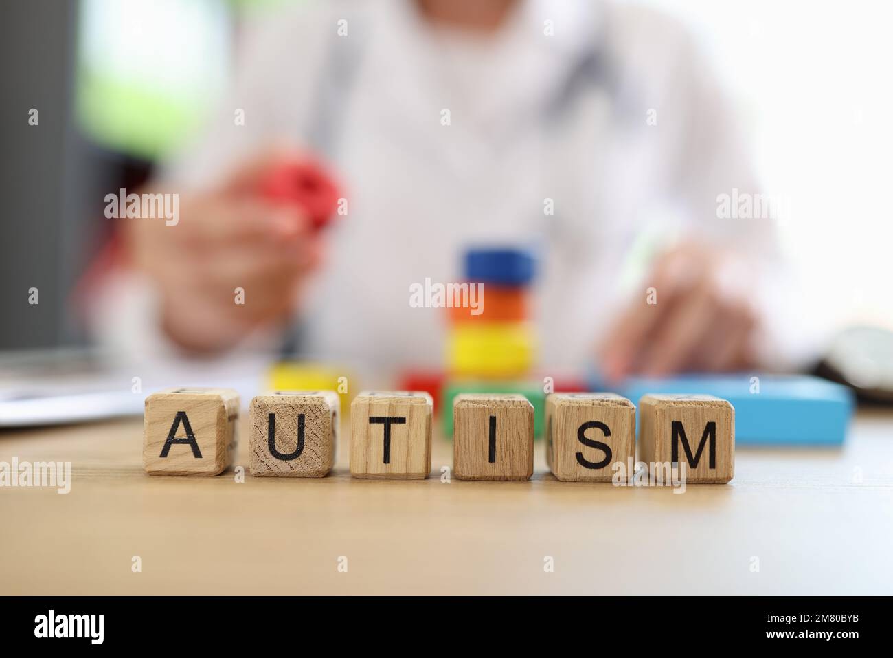Wort Autismus auf Holzblöcken aus der Nähe und verschwommener Arzt mit mehrfarbigen Bausteinen und Spielzeugen im Hintergrund. Stockfoto