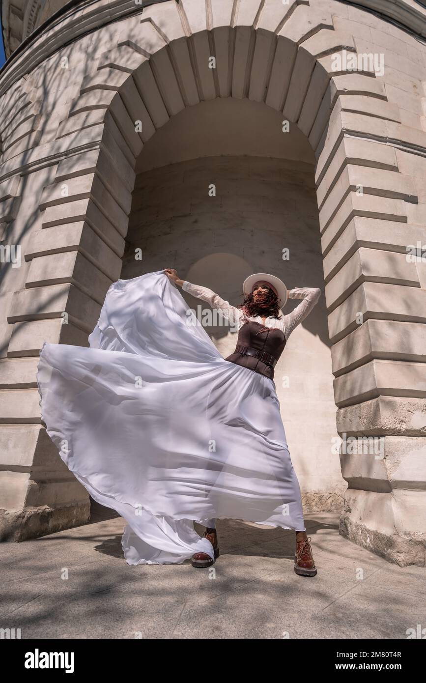 Eine stilvolle Frau, die eine Stadt baut. Eine Tänzerin in einem langen weißen Rock tanzt vor einem Gebäude mit einem Bogen. Der Rock entwickelt sich im Wind. Stockfoto