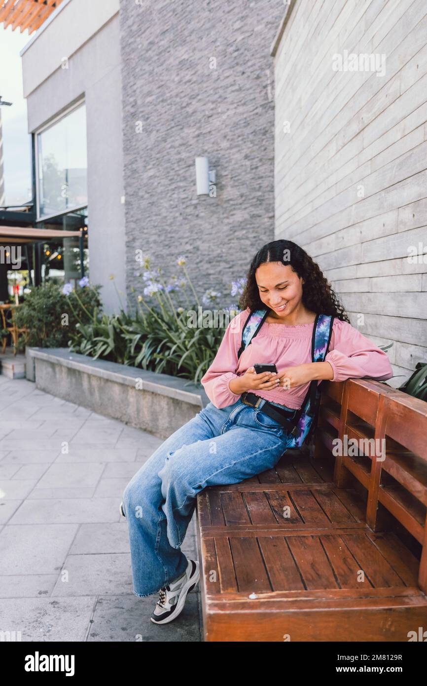 Eine entspannte schwarze Studentin, die auf einer Bank sitzt und soziale Medien auf ihrem Smartphone nutzt Stockfoto
