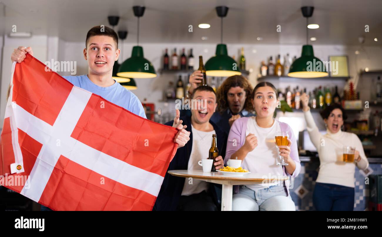 Brüllende junge Sportfans, die auf ihre Lieblingsmannschaft feuern und mit der Flagge Dänemarks wedeln, während sie gemeinsam im Pub das Spiel sehen Stockfoto