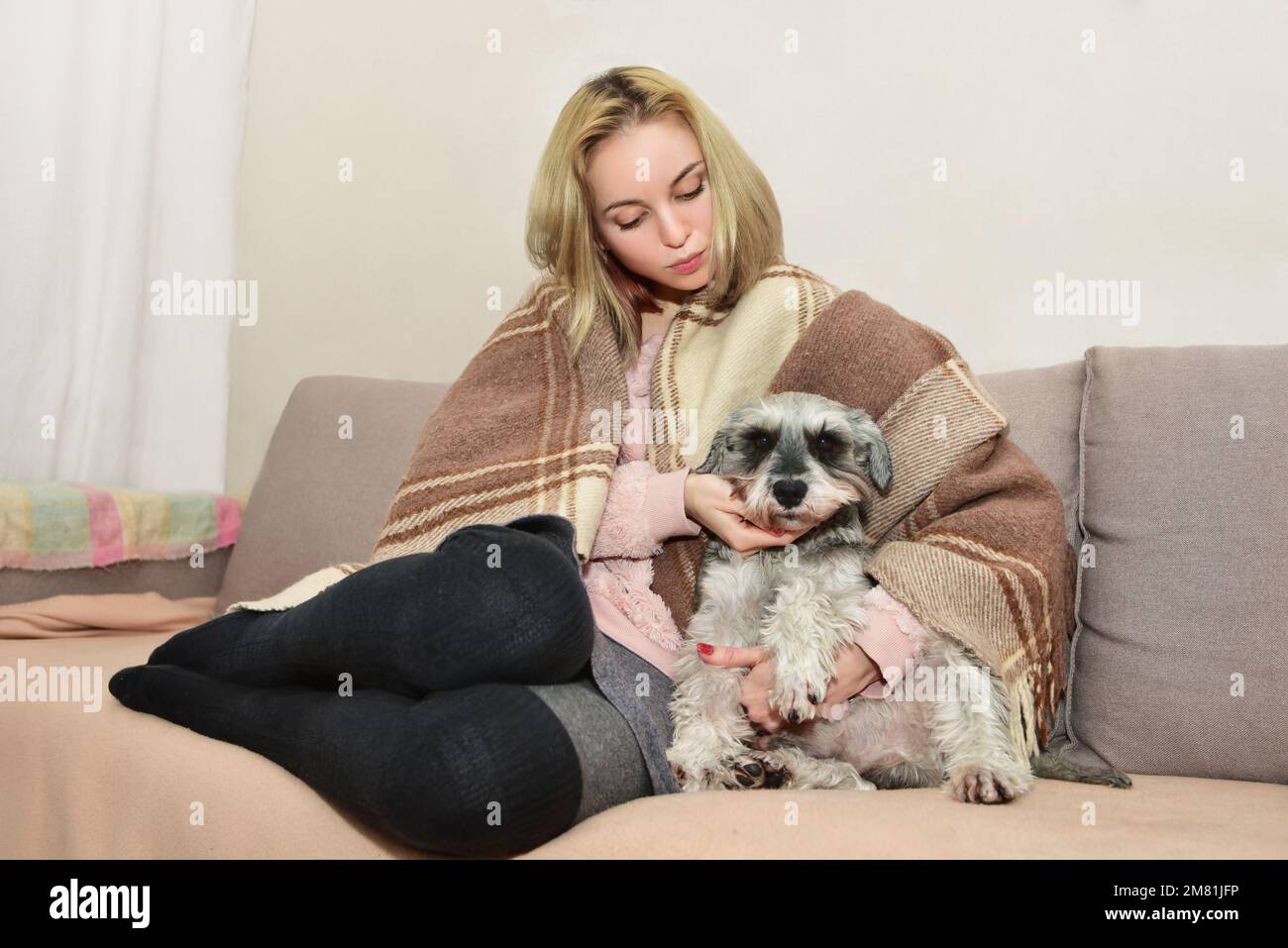 Eine wunderschöne blonde Frau in einer warmen Decke sitzt bequem auf dem Sofa und umarmt ihren süßen Hund Stockfoto