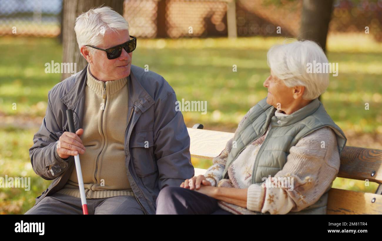 Ein blinder Senior-Mann mit Sonnenbrille ruht sich mit seiner Frau auf einer Bank im Park aus. Hochwertiges Foto Stockfoto