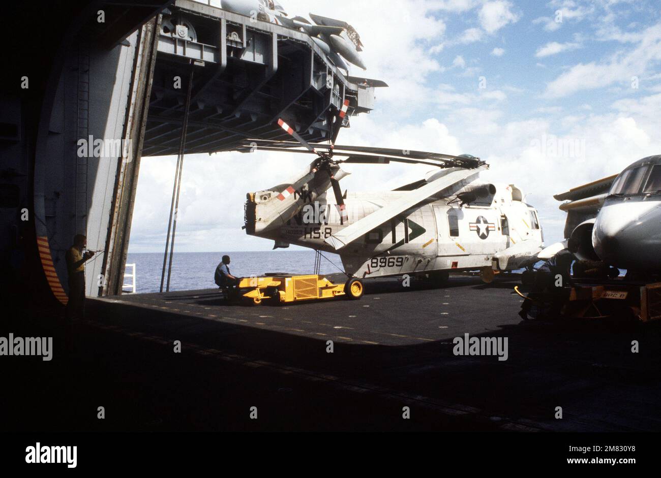Ein Sattelschlepper SD-1D wird verwendet, um einen SH-3H Sea King von Helicopter Anti-U-Boot Squadron 8 in einem abgesenkten Aufzug an Bord des Flugzeugträgers USS CONSTELLATION (CV 64) zu manövrieren. Auf der rechten Seite befindet sich ein Wikingerflugzeug S-3A. Der Spediteur nimmt an Flottenübung 85 Teil. Basis: USS Constellation (CV 64) Land: Pazifik (POC) Stockfoto