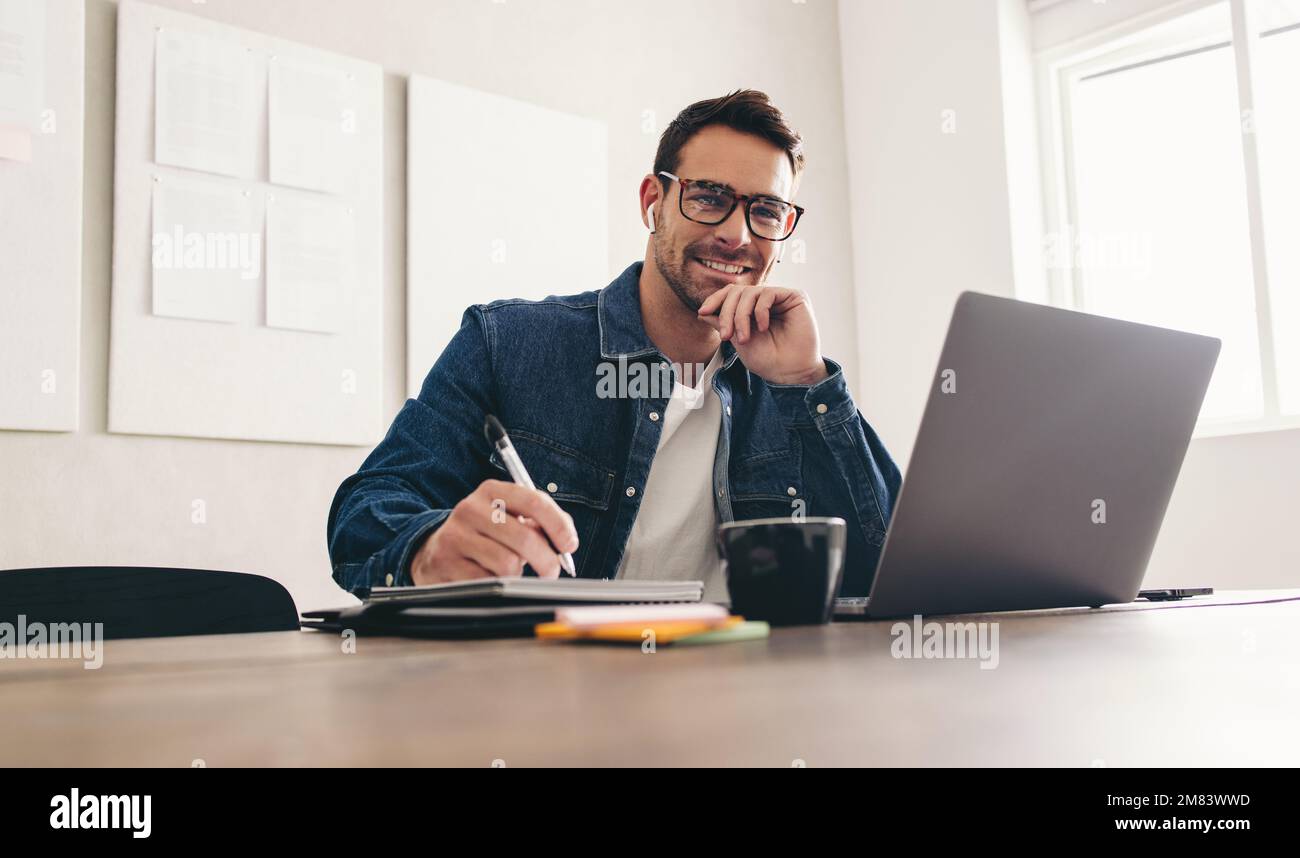 Notizen während einer Videokonferenz machen. Glücklicher junger Geschäftsmann, der in einem Online-Meeting mit einem Notebook vor der Kamera lächelt. Fröhlicher Bus Stockfoto