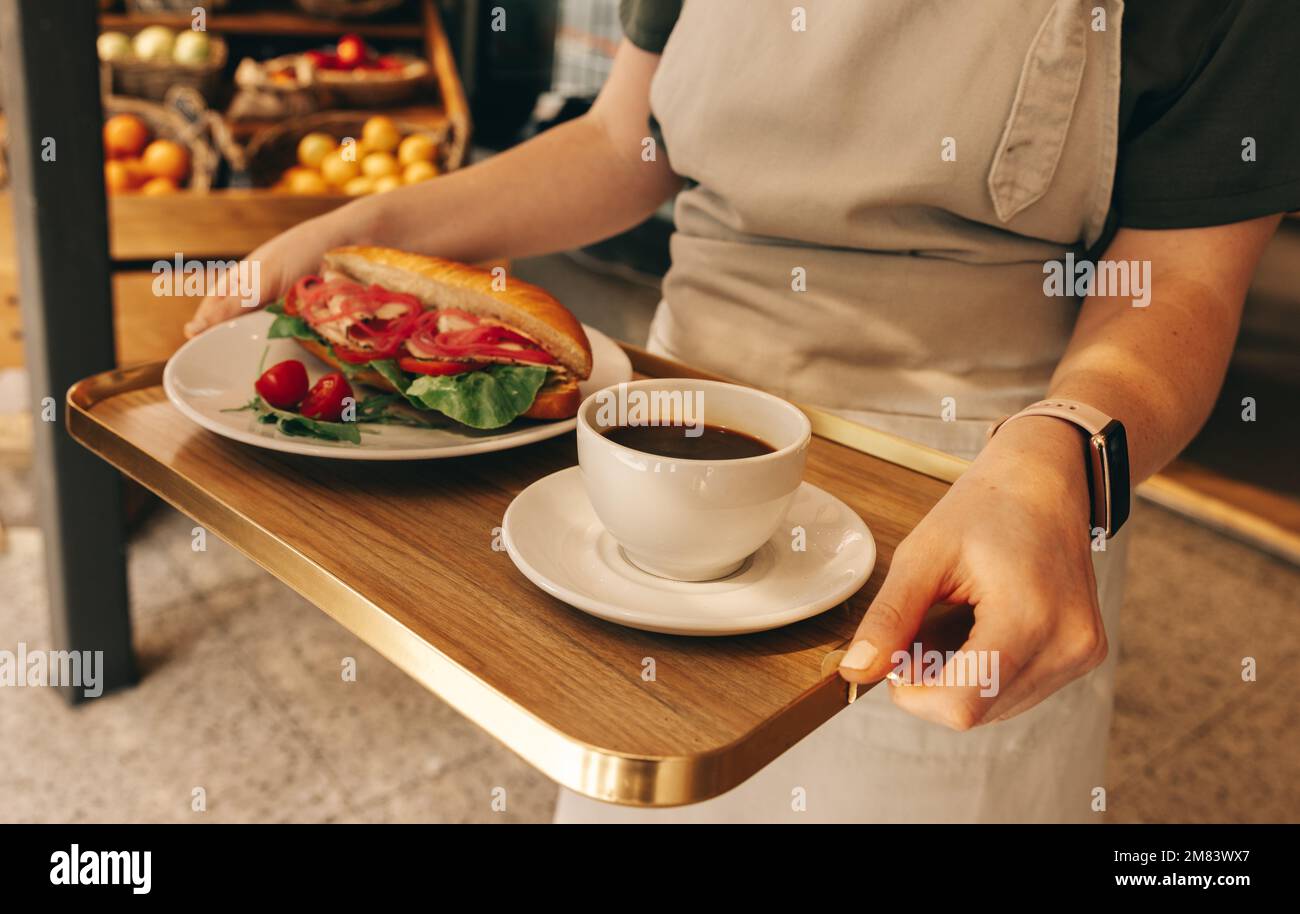Unbekannte junge Kellnerin, die ein Tablett mit einem Sandwich und einer Tasse Kaffee trug. Junge Frau, die als Kellnerin in einem Fast-Food-Restaurant arbeitet. Stockfoto