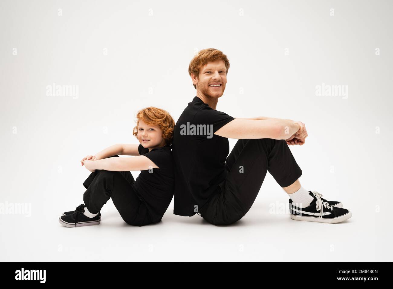 Die ganze Länge voller fröhlicher rothaariger Vater und Sohn in schwarzen T-Shirts und Hosen, die Rücken an Rücken auf hellgrauem Hintergrund sitzen, Stockbild Stockfoto