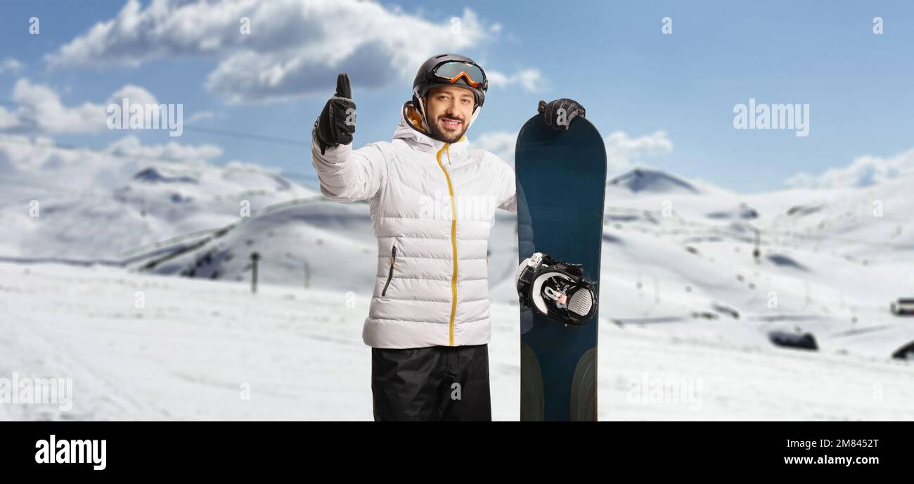 Ein cooler junger Mann auf einem Berg mit einem Snowboard, das Daumen nach oben zeigt Stockfoto