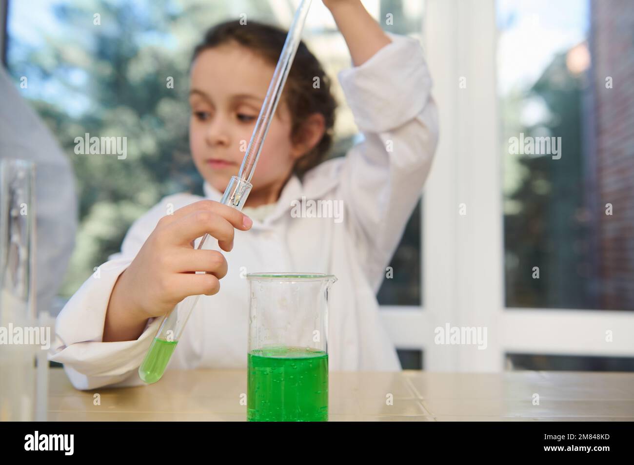 Details: Messpipette und Reagenzglas mit chemischer Flüssigkeit in den Händen eines verschwommenen Grundschulmädchens. Chemieklasse Stockfoto