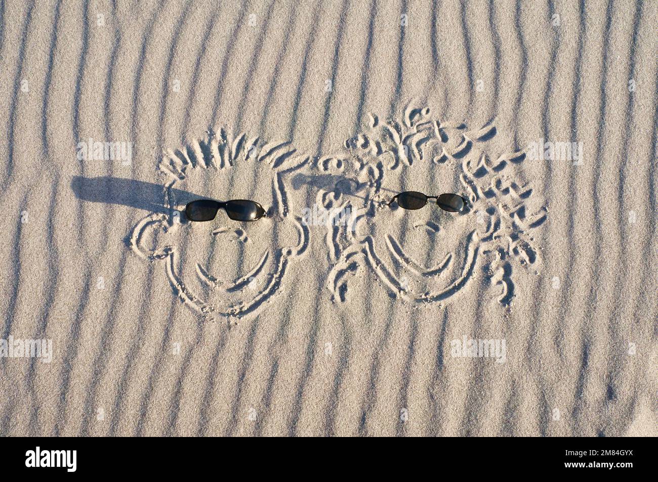 Gesicht im Sand am Strand gezeichnet, mit Sonnenbrille. Sand mit Wellenmuster. Ururlaubsfoto vom Meer Stockfoto