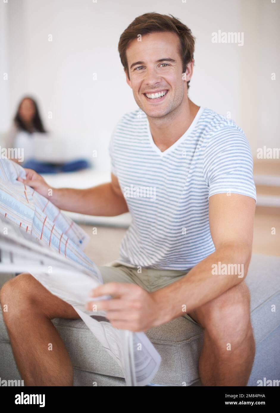 Ich lese die Sonntagszeitungen. Ein junger Mann, der lächelt und eine Zeitung hält, während seine Freundin im Hintergrund sitzt. Stockfoto