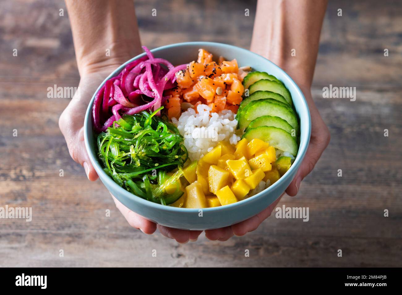 Frauenhände halten eine Schüssel mit Reis, Lachs, Gurke, Mango, Zwiebeln, Wachsalat, Mohnsamen und Sonnenblumenkernen auf Holzhintergrund Stockfoto