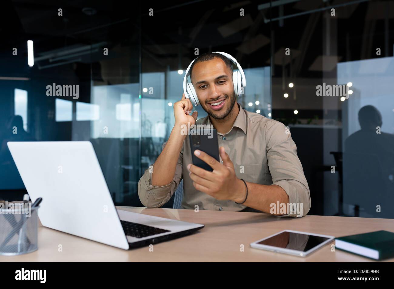 Erfolgreicher Geschäftsmann am Arbeitsplatz, der Online-Videos ansieht und Musik, Hörbücher und Podcasts anhört, hispanischer Mann im Büro, der an einem Laptop mit Smartphone und Kopfhörern arbeitet. Stockfoto