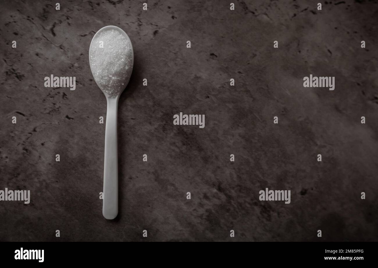 Ein Löffel mit weißem raffiniertem Zucker, isoliert auf einer dunklen Küchentheke. Draufsicht flach liegend mit freiem Platz für Text auf der rechten Seite Stockfoto