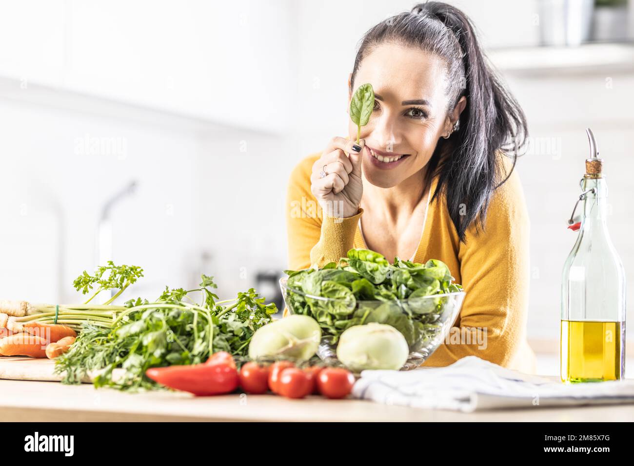 Nette junge Frau, die ein Spinatblatt hält, ihr Auge bedeckt, lächelt, frisches Gemüse auf dem Tisch neben ihr. Stockfoto