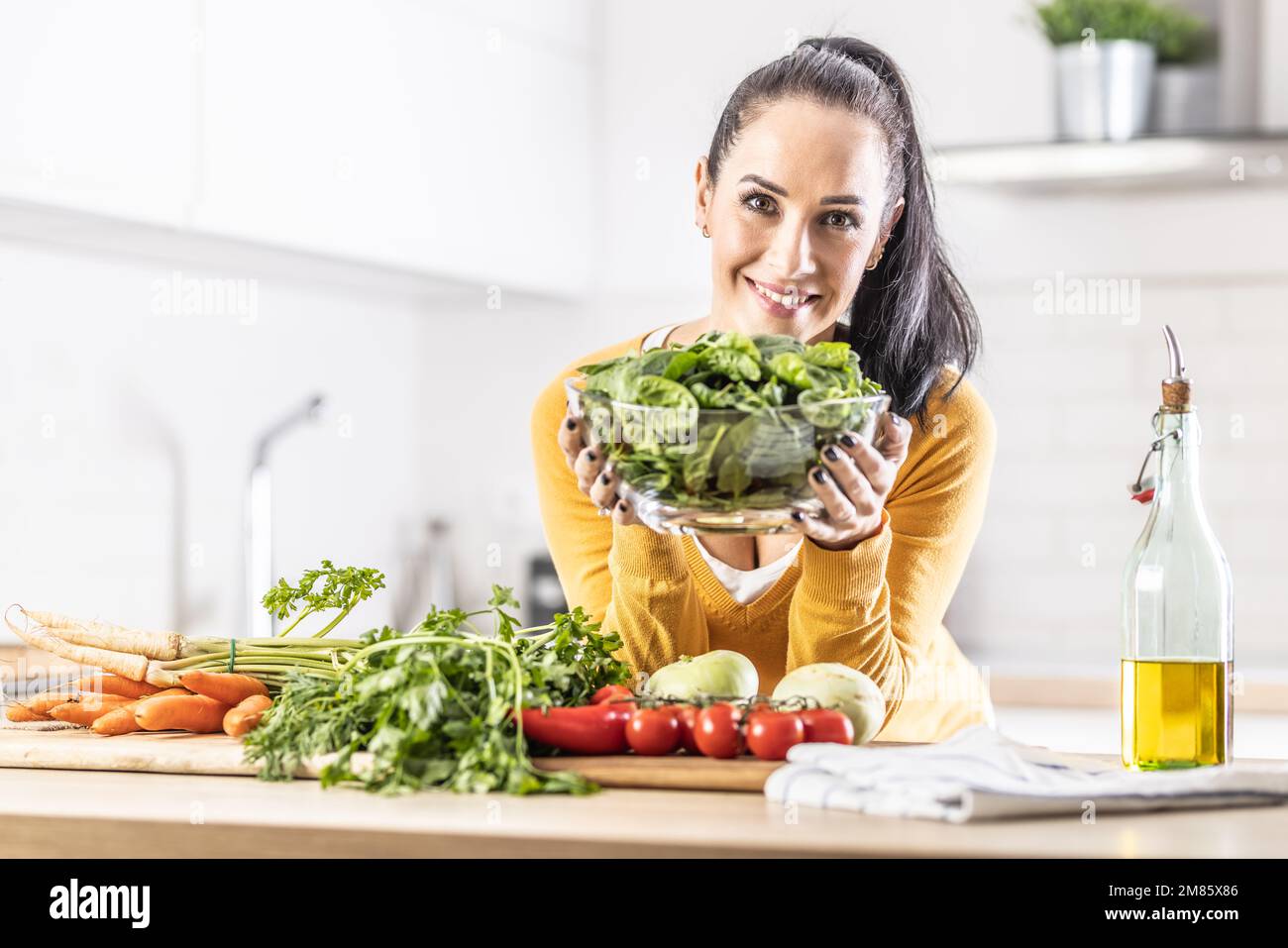 Lächelnde Frau mit einer Schüssel voller Spinatblätter zusammen mit frischem Gemüse und Olivenöl in ihrer Küche. Stockfoto