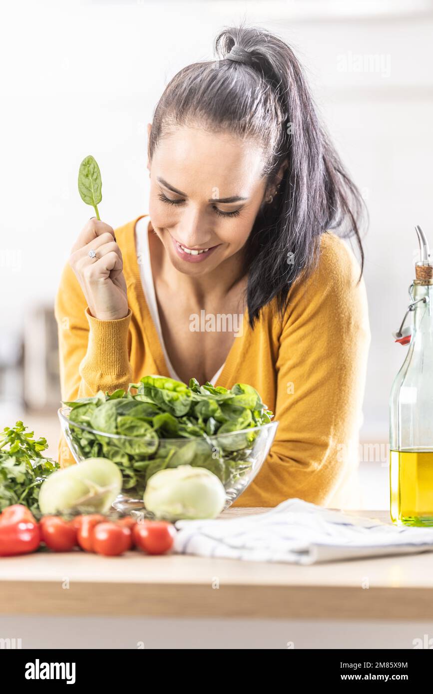 Nette junge Frau, die ein Spinatblatt hält, lächelt, frisches Gemüse auf dem Tisch neben ihr. Stockfoto