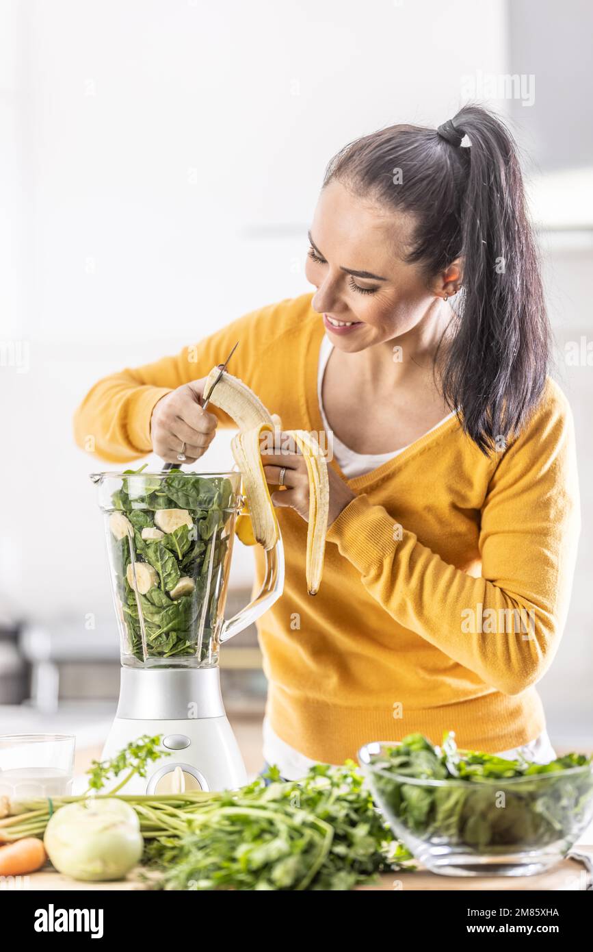 Eine Frau in der Küche schneidet eine Banane in einem Mixer zusammen mit Spinat und macht einen gesunden Smoothie. Stockfoto