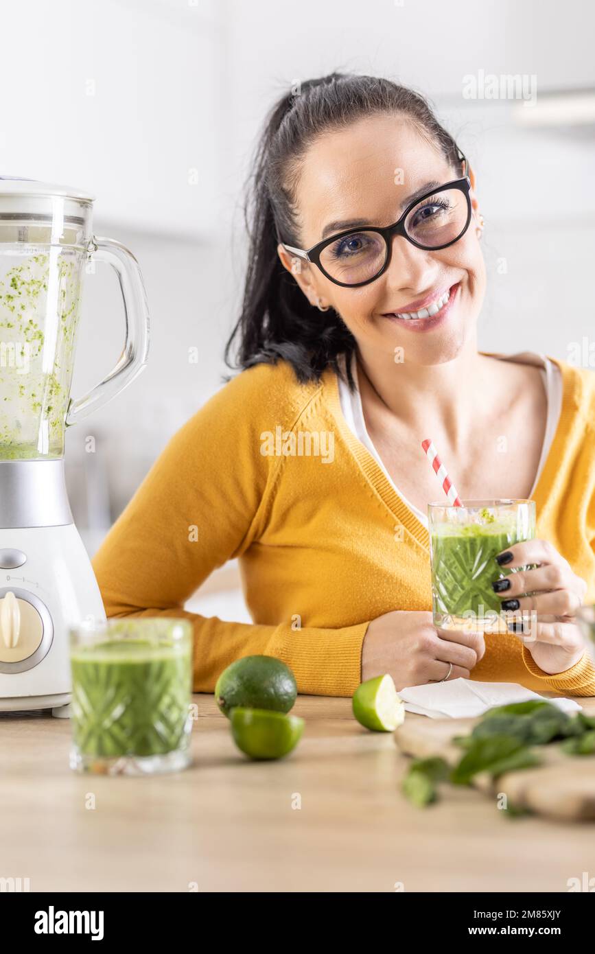 Glückliche Frau mit Spinat-Smoothie-Getränk oder vegetarischem Shake, die in ihrer Küche sitzt. Stockfoto