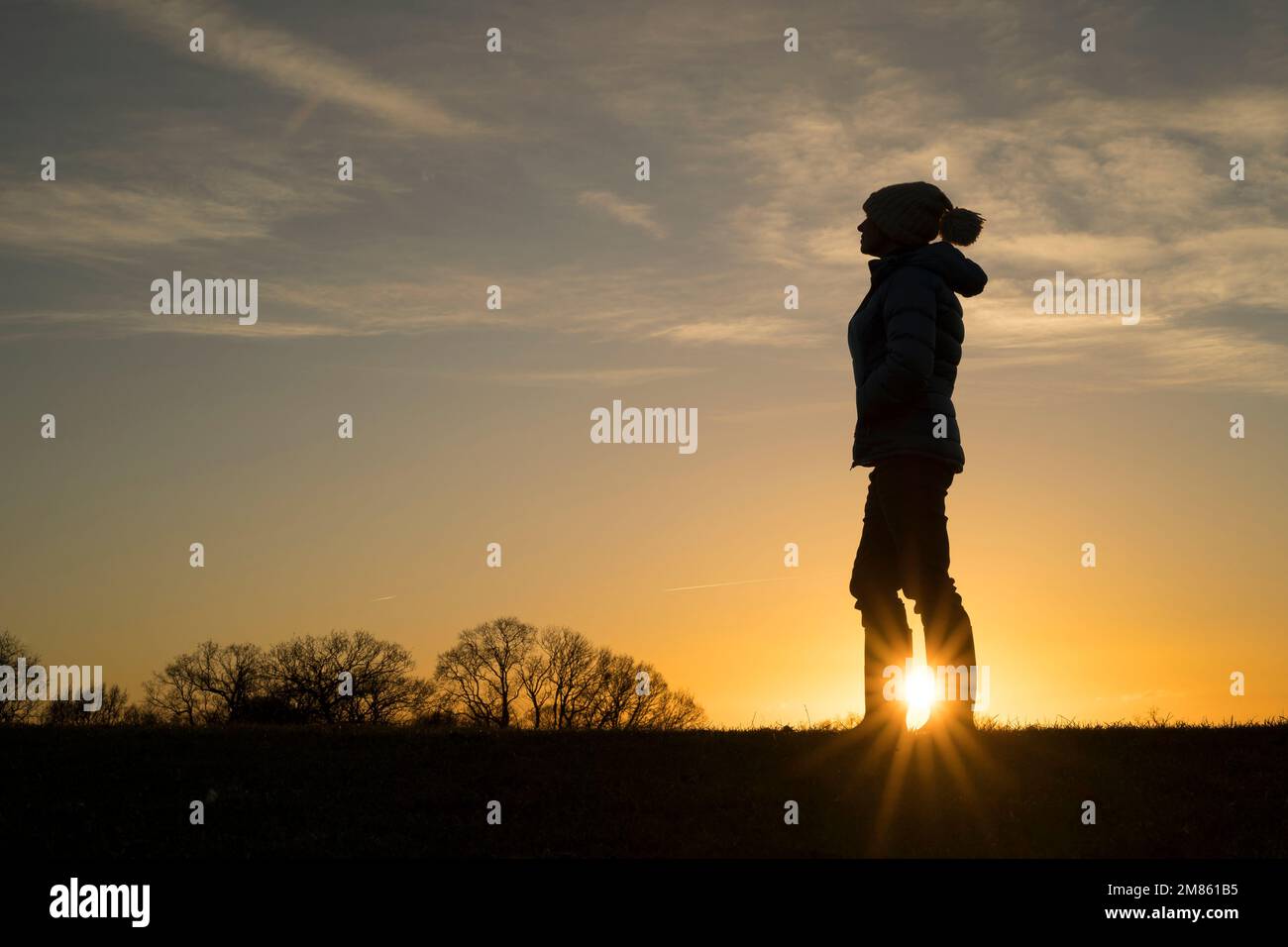 Ein Sonnenschein bricht durch die Silhouette einer isolierten Frau, die in einem warmen Mantel und einem Hut gekleidet ist, an einem späten Nachmittag, einem Winterspaziergang durch die Landschaft Großbritanniens. Stockfoto