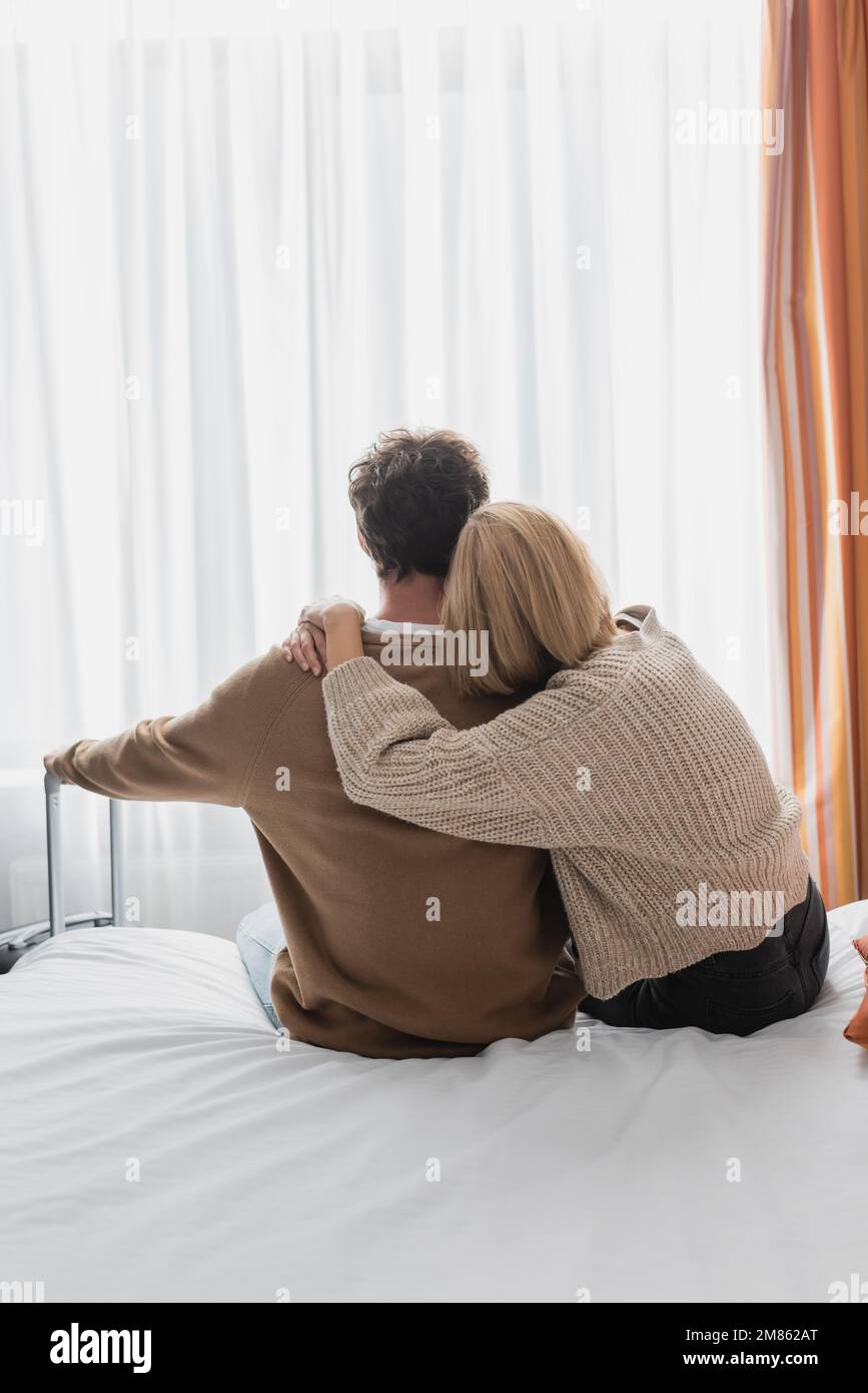 Rückansicht einer blonden Frau, die ihren Freund umarmt und auf dem Bett im Hotel neben dem Koffer sitzt, Stockbild Stockfoto