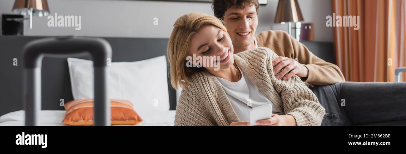 Glückliche blonde Frau, die sich mit ihrem Handy in der Nähe ihres Freundes im Bett im Hotel unterhielt, Banner, Stock Image Stockfoto