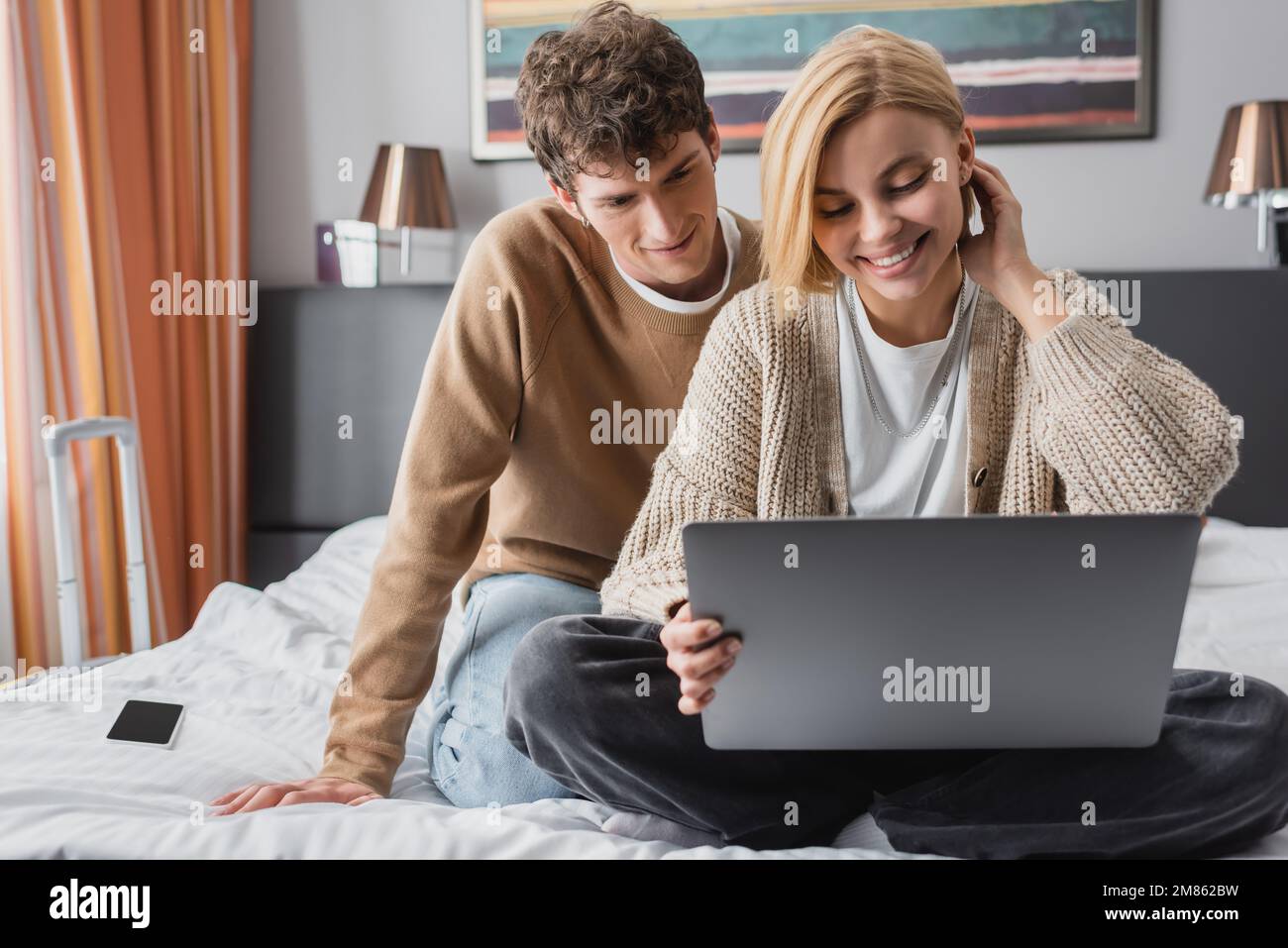 Lächelnde blonde Frau, die mit einem Laptop auf dem Hotelbett neben ihrem jungen Freund sitzt, Bild der Börse Stockfoto