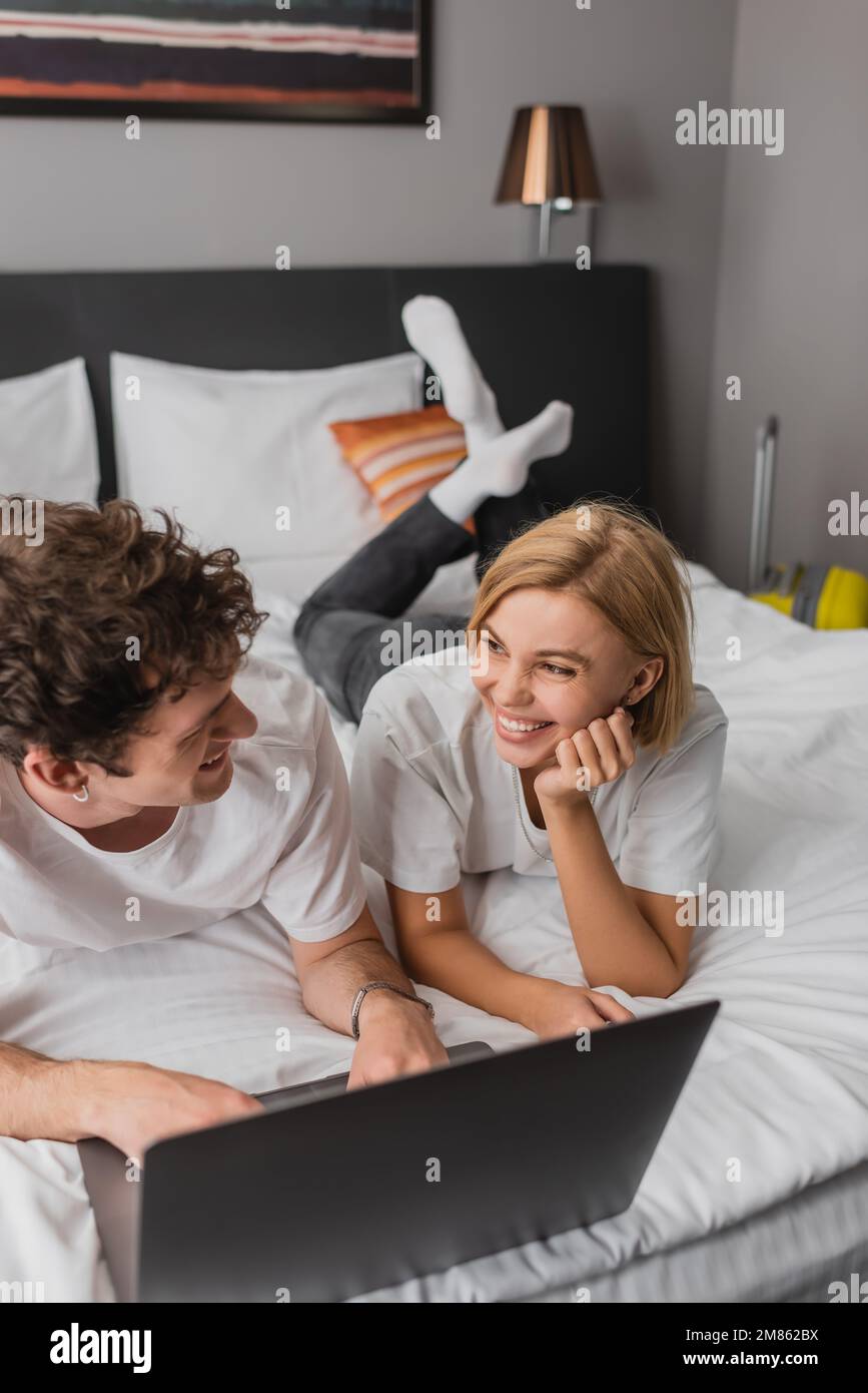 Fröhliche blonde Frau, die ihren Freund in der Nähe des Laptops im Hotel ansieht, Bild der Aktie Stockfoto