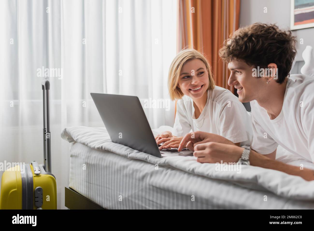 Glückliche blonde Frau, die ihren Freund in der Nähe des Laptops und der Reisetasche im Hotelschlafzimmer anschaute, Bild der Aktien Stockfoto