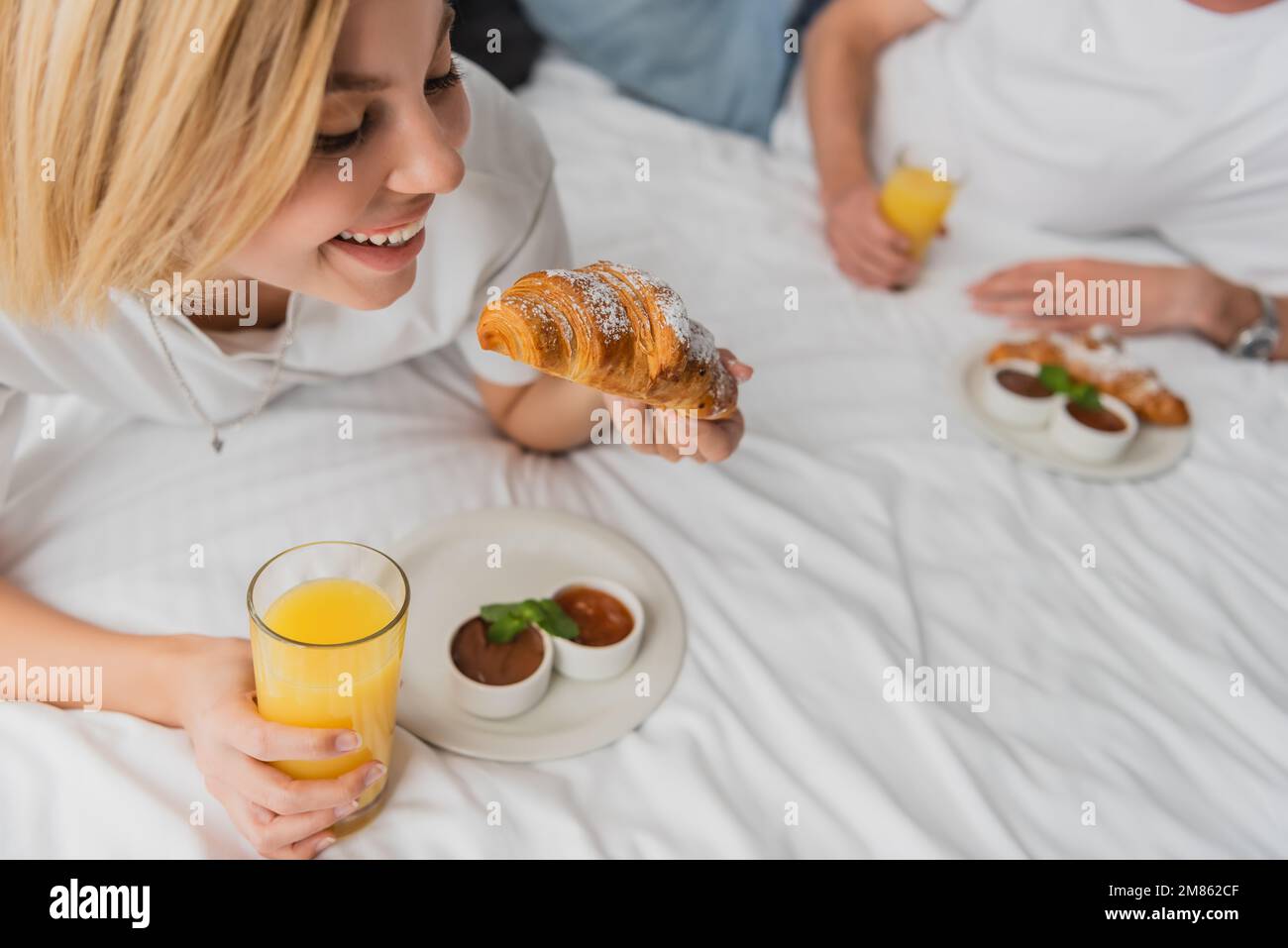 Eine glückliche blonde Frau, die leckere Croissants und Orangensaft in der Hand hielt, während sie im Hotelbett frühstückte, in der Nähe eines verschwommenen Freundes, Bild der Aktien Stockfoto