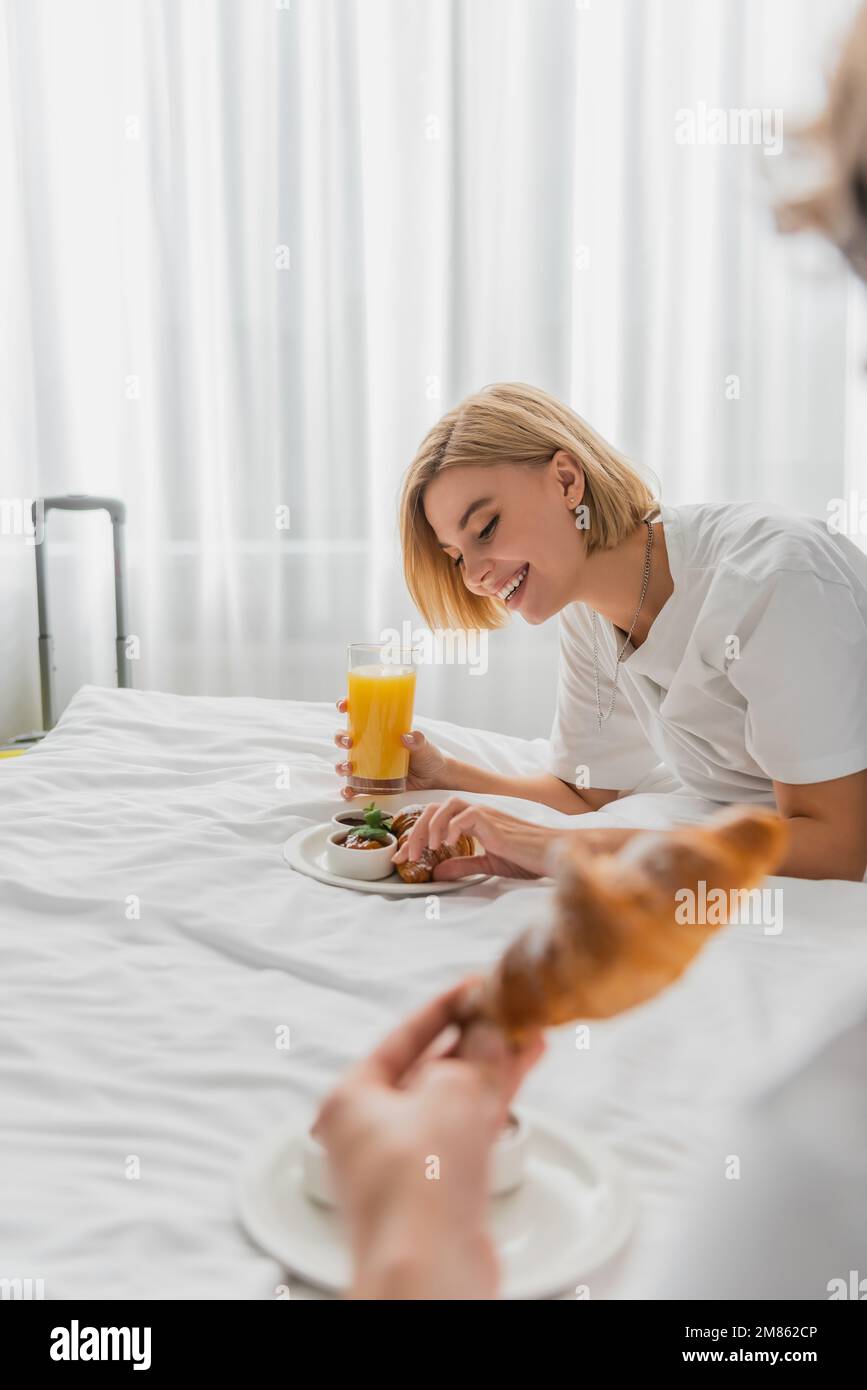 Eine fröhliche blonde Frau, die Frühstück mit Orangensaft und Croissant auf dem Hotelbett hatte, in der Nähe eines verschwommenen Freundes, Bild der Aktien Stockfoto