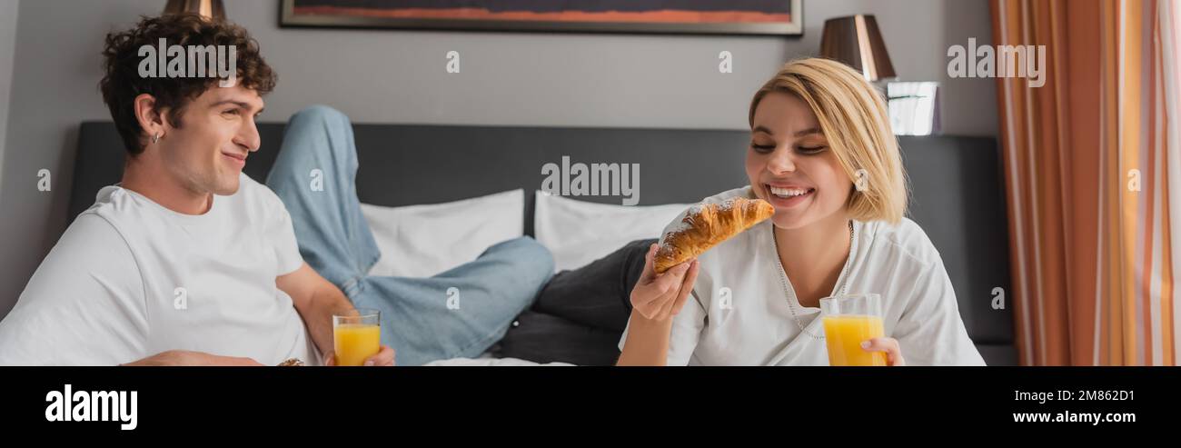 Eine glückliche blonde Frau hielt leckere Croissants und Orangensaft in der Nähe eines lächelnden Freundes im Hotelzimmer, Banner, Stockbild Stockfoto