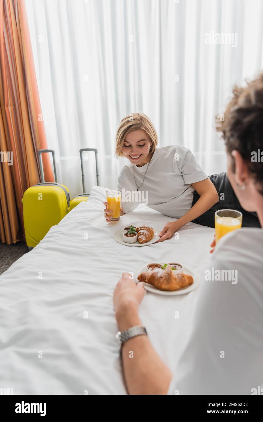 Eine glückliche blonde Frau, die Orangensaft und einen Teller mit Croissant in der Hand hatte, fast ein verschwommener Freund im Hotelzimmer, Bild der Aktien Stockfoto