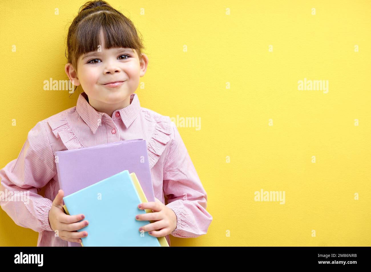 Fleißiges Kind, das Bücher in der Hand hält und isoliert auf gelben Hintergrund in die Kamera schaut. Das kleine Kind liebt Bildung, Lernen Stockfoto