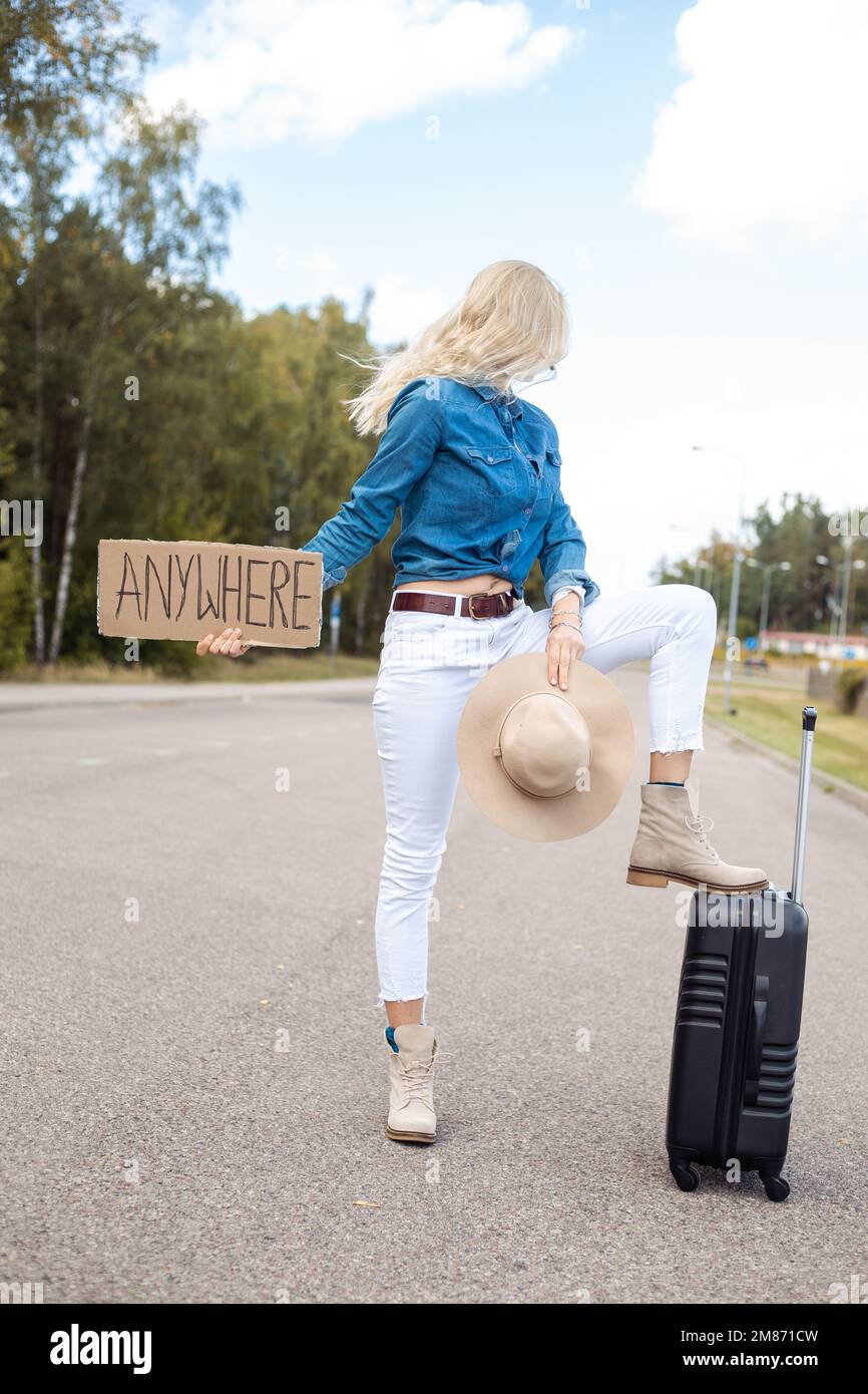 Eine Frau mit Koffer und Pappposter hält an einem leeren Highway vorbei und schaut zurück. Lady mit Hut, entfliehen Sie der Stadt, um überall hinzugehen. Auf Reisen Stockfoto