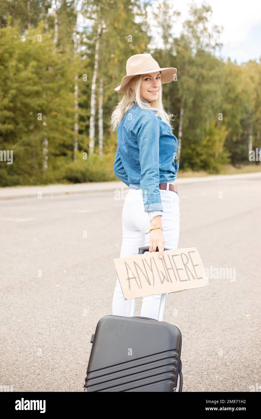 Glückliche Frau ging auf einer leeren Straße mit Pappposter und Koffer, Rückansicht. Lady mit Hut und Jeansanzug Flucht aus der Stadt per Auto Stop zum Mitnehmen Stockfoto