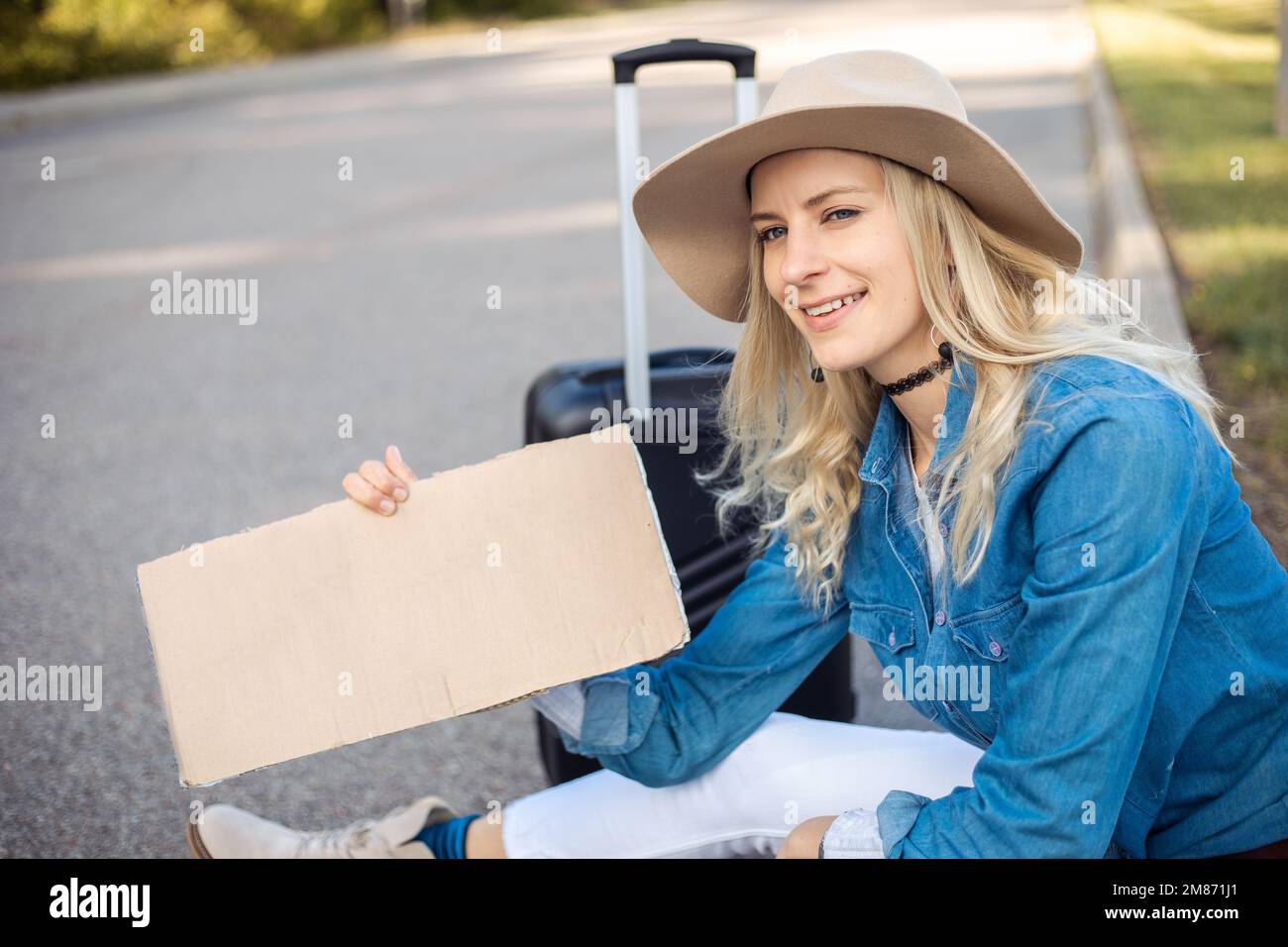 Glückliche Frau, die hoffentlich am Auto vorbeifährt, sitzt mit Koffer und Pappposter am Straßenrand einer leeren Straße. Lady entfliehen aus der Stadt, um überall hinzugehen Stockfoto