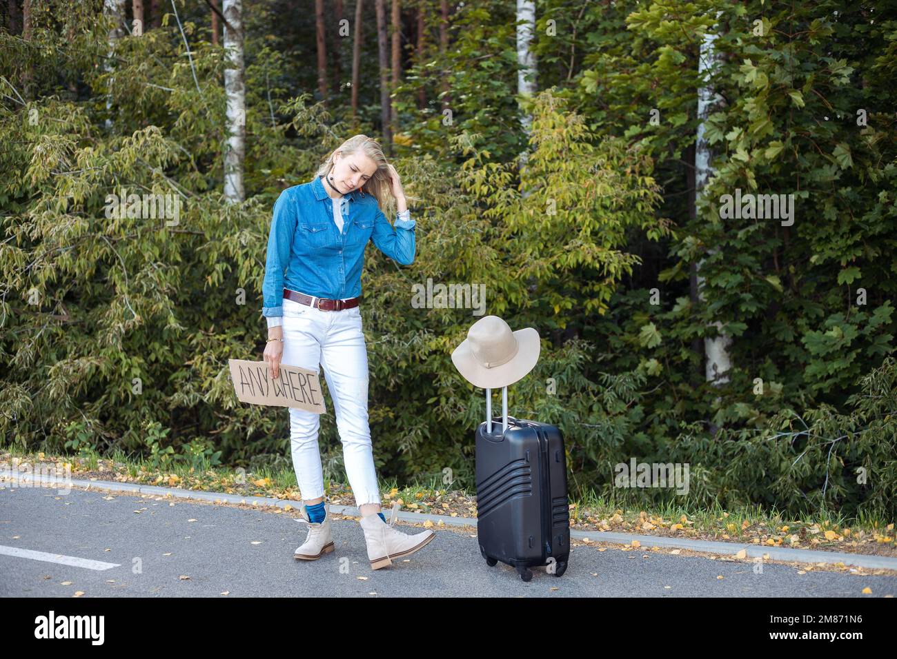 Eine junge Frau wartet im Wald am Auto vorbei und steht mit Koffer und Pappposter am Straßenrand. Reisender fliehen aus der Stadt, um überall hin zu kommen. Auf Reisen Stockfoto
