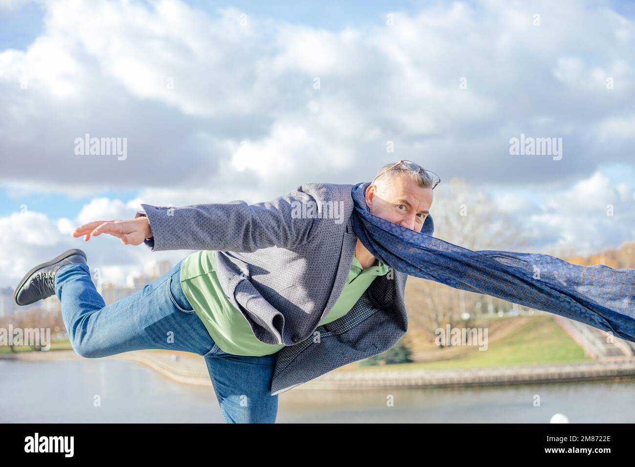 Ein lustiger, reifer, grilliger Mann in warmer Kleidung steht an einem windigen Tag im Park auf einem Bein mit erhobenen Beinen und Armen an der Grenze Stockfoto