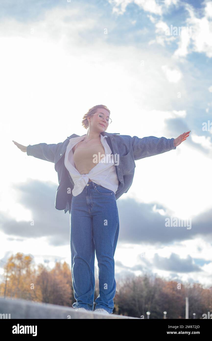 Ruhige, zufriedene, spirituelle Frau in Jeans Outfit steht an der Grenze mit erhobenen Armen wie in der Titanic gegen sonnigen bewölkten Himmel Stockfoto