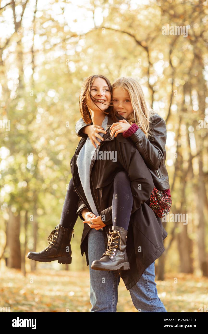 Vertikale verspielte, positive, liebevolle, fröhliche Familie, junge Mutter hält Tochter auf dem Rücken, Promenade im goldenen Herbstwald Stockfoto