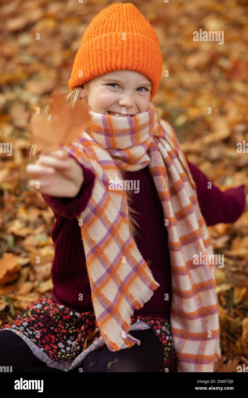 Senkrecht lächelnd positiv fröhlich, fröhlich, entzückend, lächelndes kleines blondes Mädchen mit orangefarbenem, warmem Hut und Pullover Stockfoto