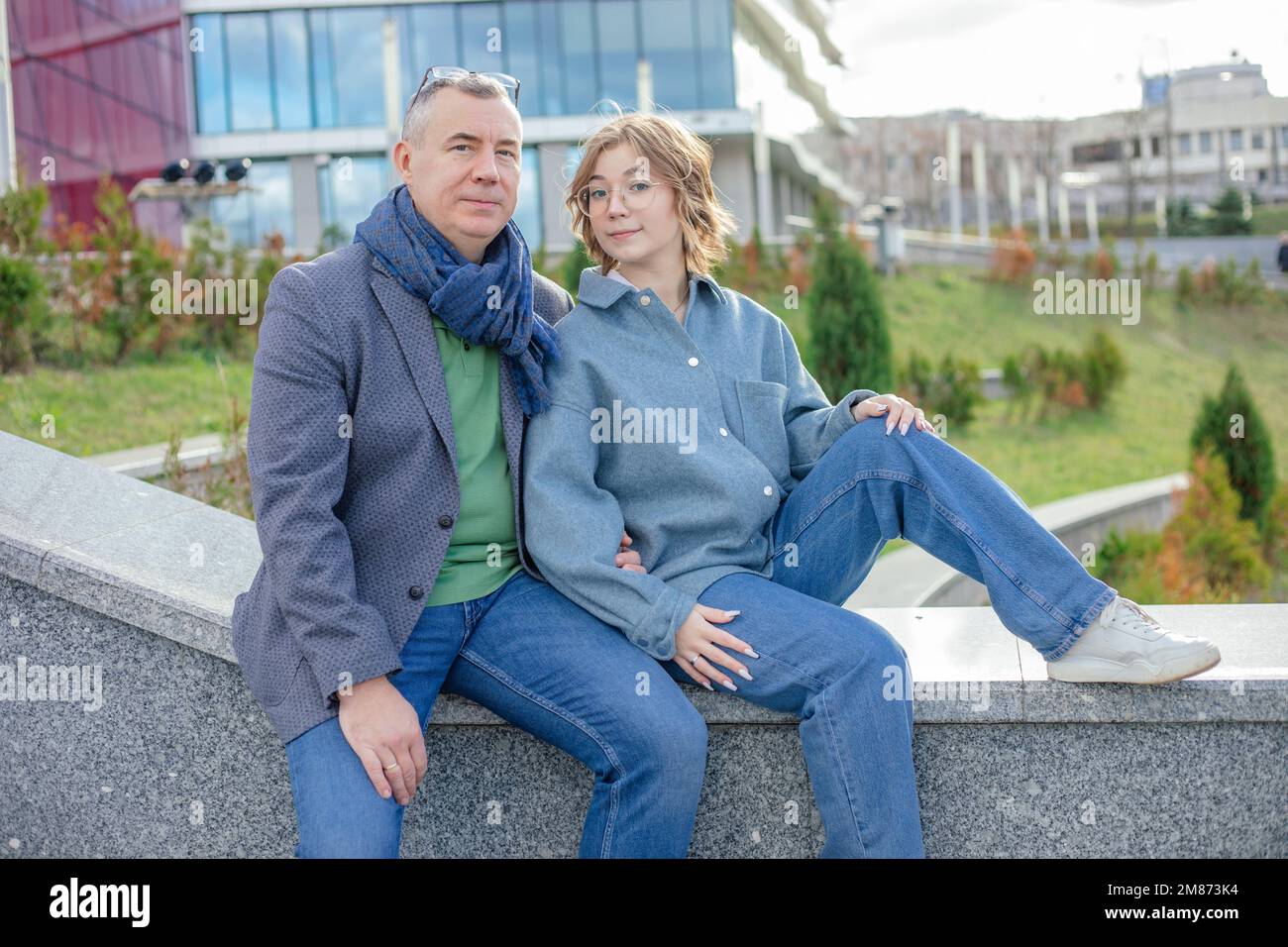 Netter, netter, lächelnder, reifer Mann, Frau in Jeans-Outfit, sitzt auf einem Steingeländer und steht vor dem Universitätsgebäude. Unterstützung des Vaters Stockfoto