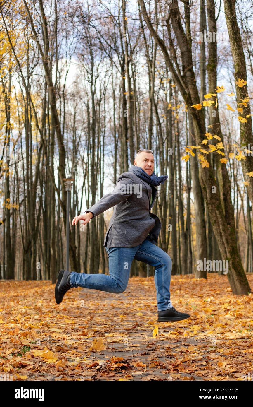 Vertikal lustig, erregter, reifer, gegrillter Geschäftsmann springt auf ein Bein mit erhobenen Beinen und Armen und schwebt im Park Stockfoto