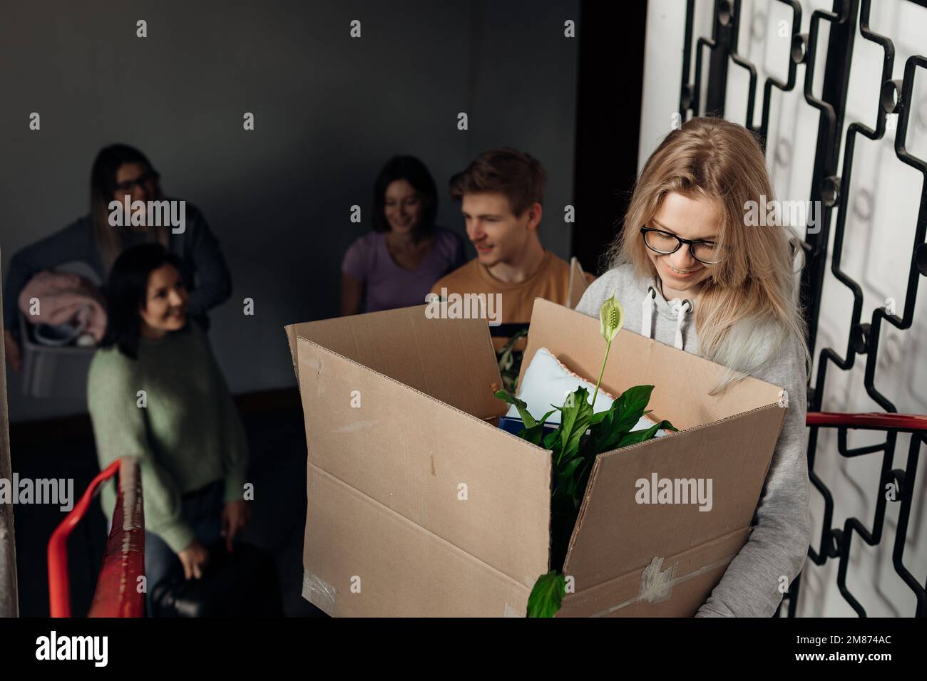 Lächelnde, fröhliche, sprechende Studenten, die in eine Unterkunft ziehen, Kisten mit Hauspflanzenzeug auf der Treppe mitnehmen und tragen Stockfoto