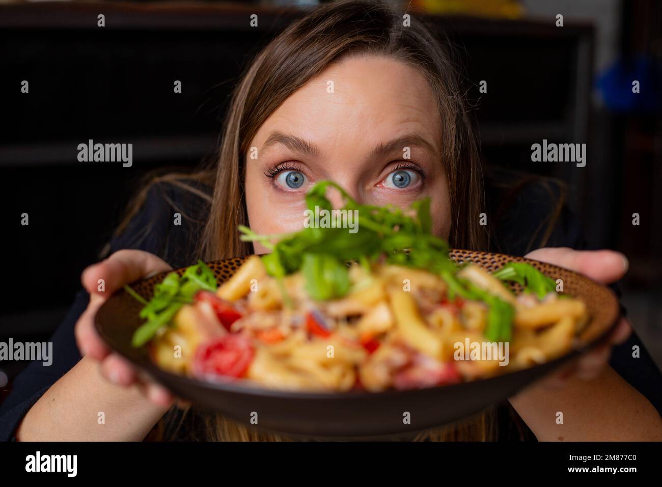 Porträt einer Frau mittleren Alters, die mit breiten Augen in die Kamera starrt und leckere Pasta mit Kirschtomaten, Rucola und Käse zeigt. Stockfoto