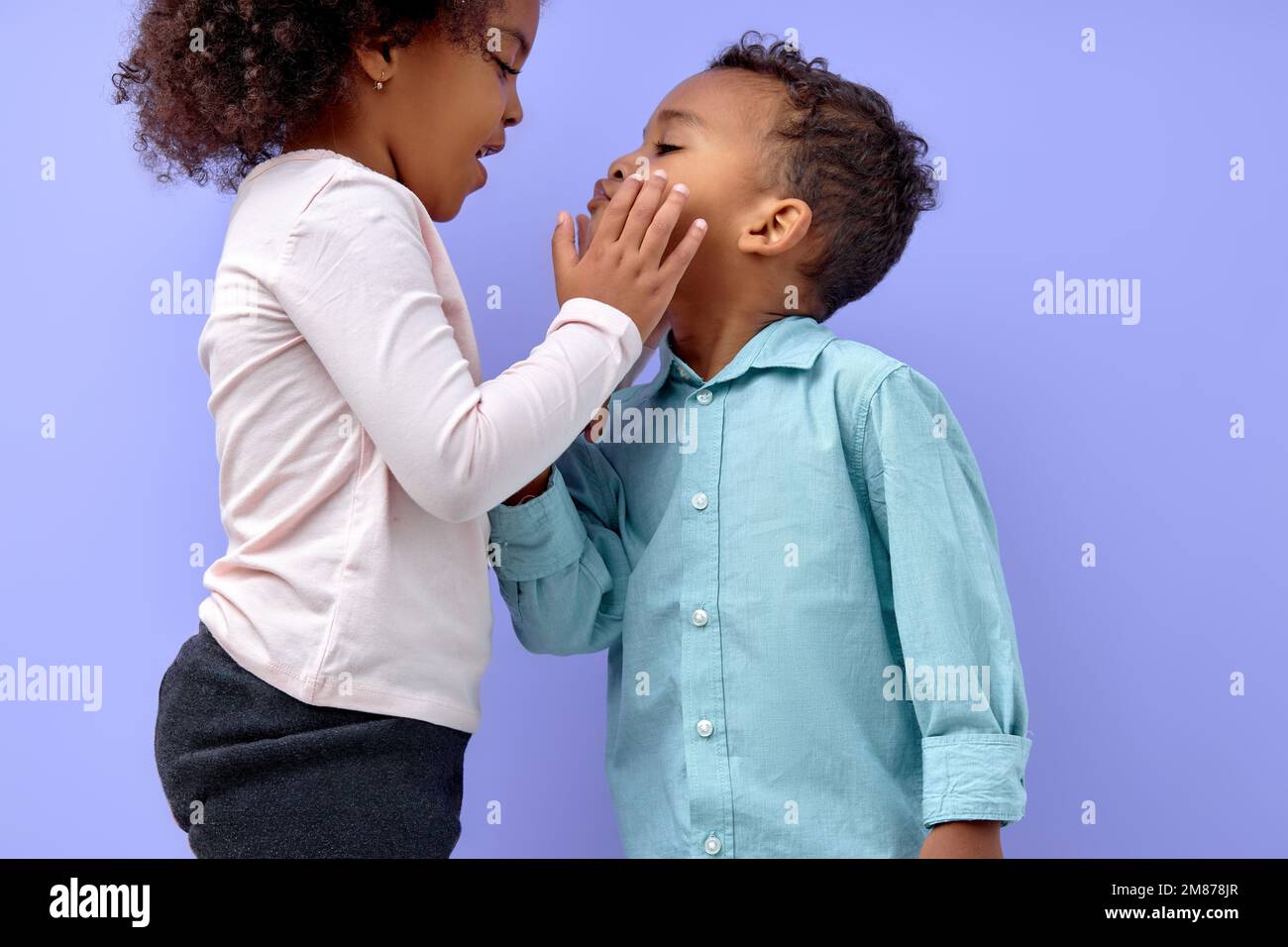 Zwei fröhliche schwarze amerikanische Geschwister, Schwester küsst ihren Bruder, isoliert auf lila Hintergrund. Porträt süßer Kinder in lässiger Kleidung. Junge und Mädchen Stockfoto
