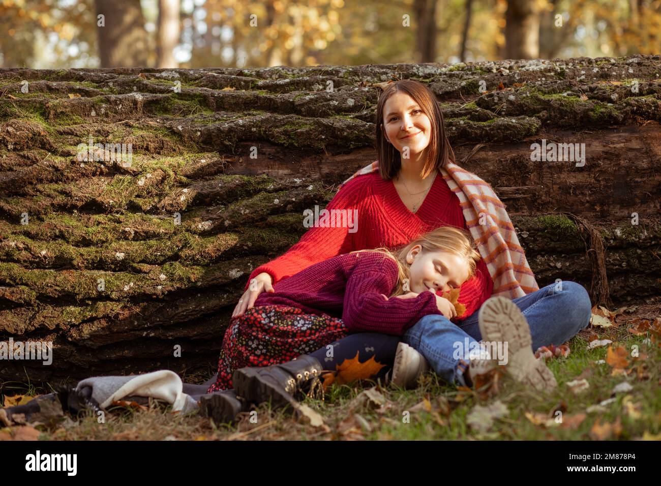 Glückliche, glückliche, entspannte, lächelnde Frau, schau auf die Kamera, halte das schlafende Mädchen auf Knien im Wald. Aktiver Lebensstil, Wandern Stockfoto