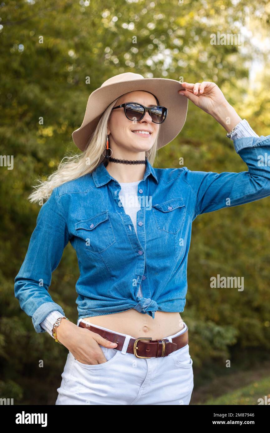 Senkrecht lächelnde, moderne, selbstbewusste, freudige, sorgenfreie blonde Frau mit Sonnenbrille und Hut, die auf die Sonne blickt und spaziert Stockfoto