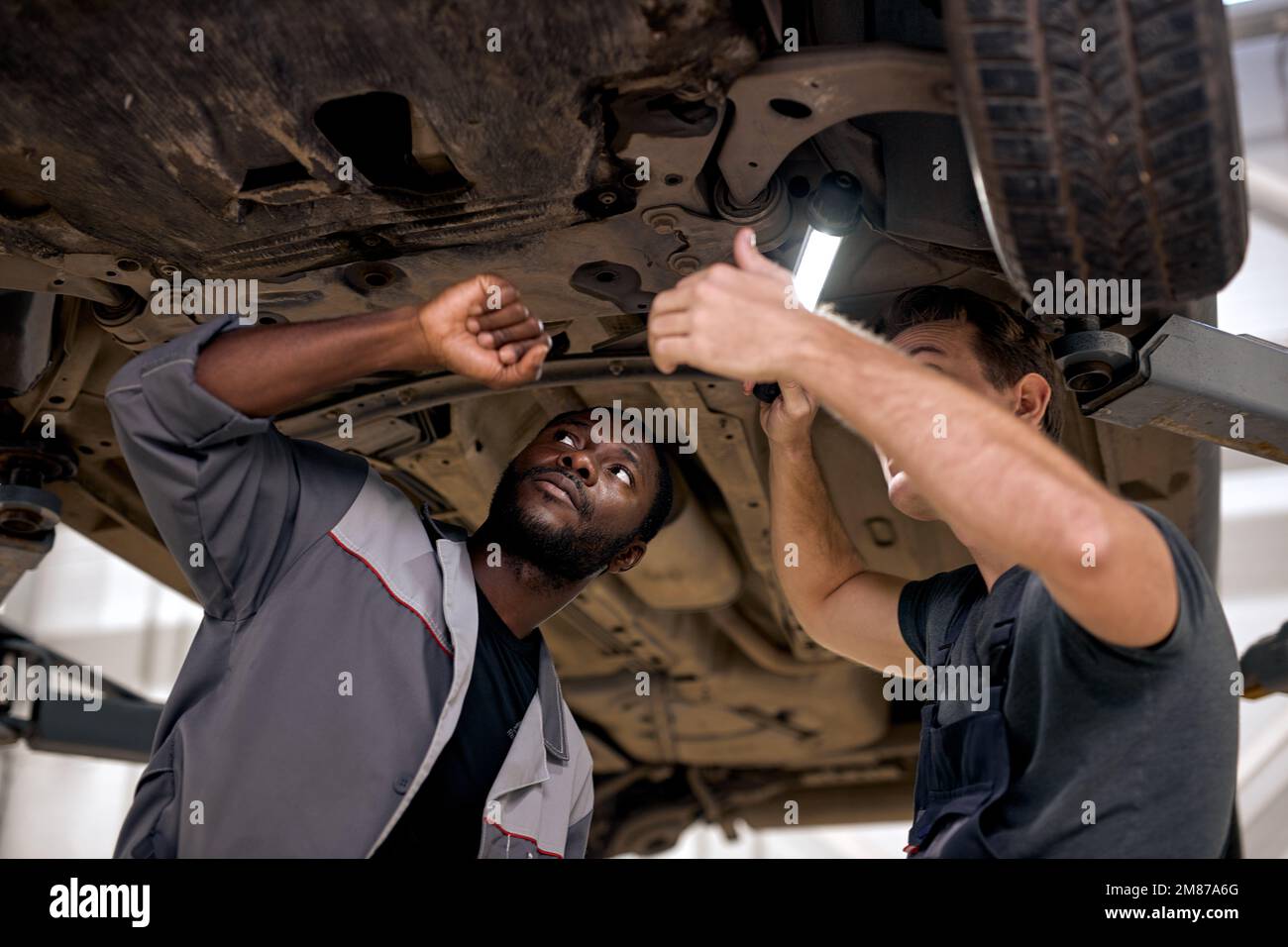 Zwei vielseitige, gutaussehende Mechaniker in Uniform untersuchen das Auto bei der Arbeit im Autoservice. Junge Männer in Overalls konzentrierten sich auf die Zusammenarbeit mit Lampen Stockfoto